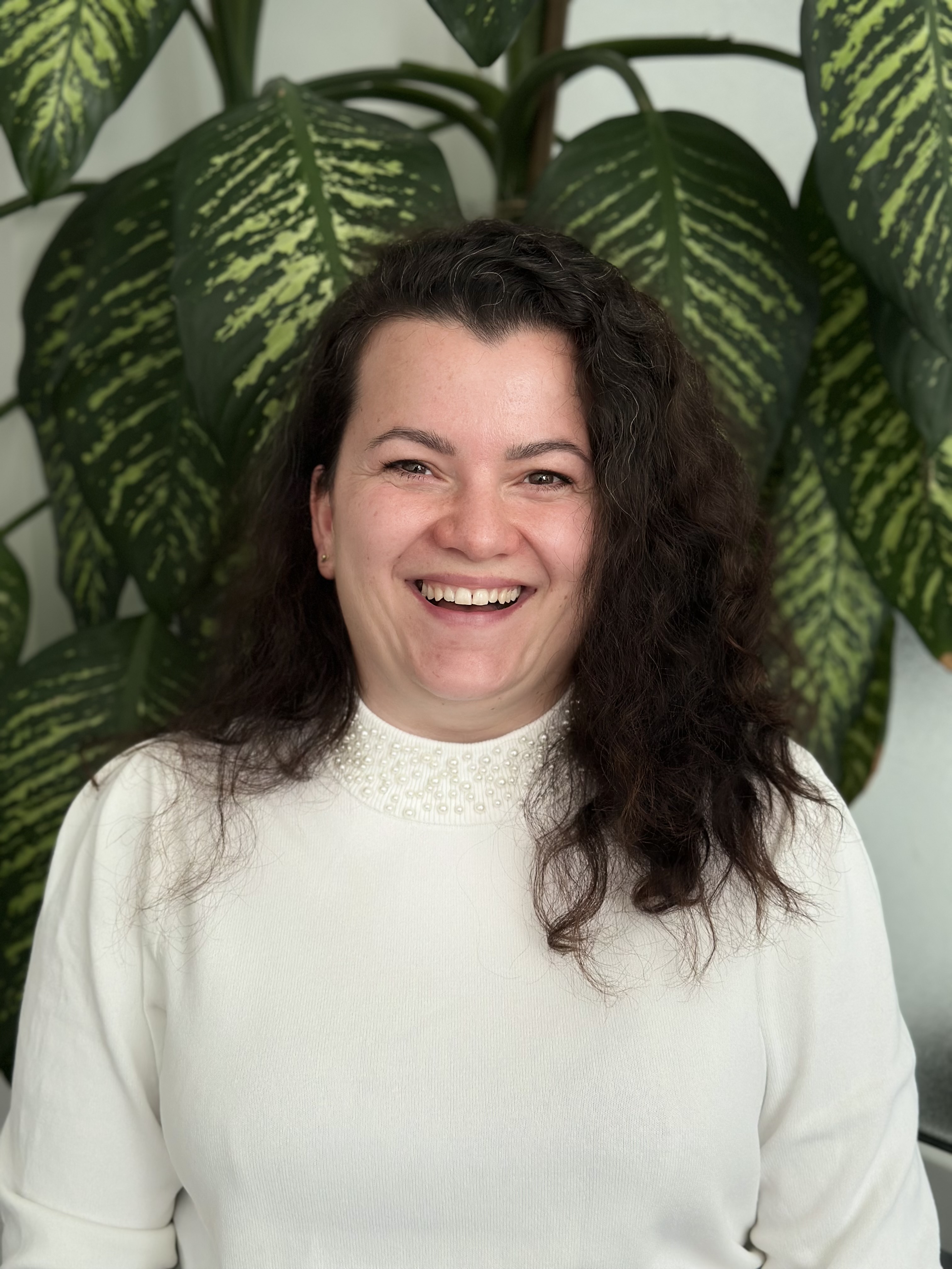 Smiling woman with curly dark hair wearing a white sweater with pearl details around the collar, standing in front of large green leaves.